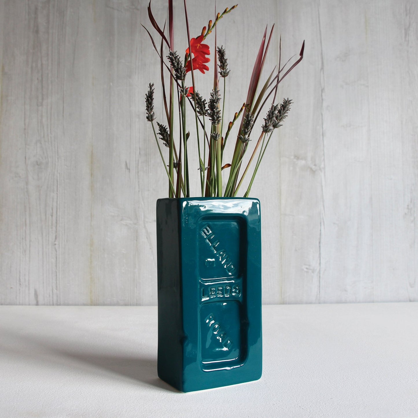 A teal ceramic brick-shaped vase with 'Leeds Elland Road' embossed on the side and flowers in the top.