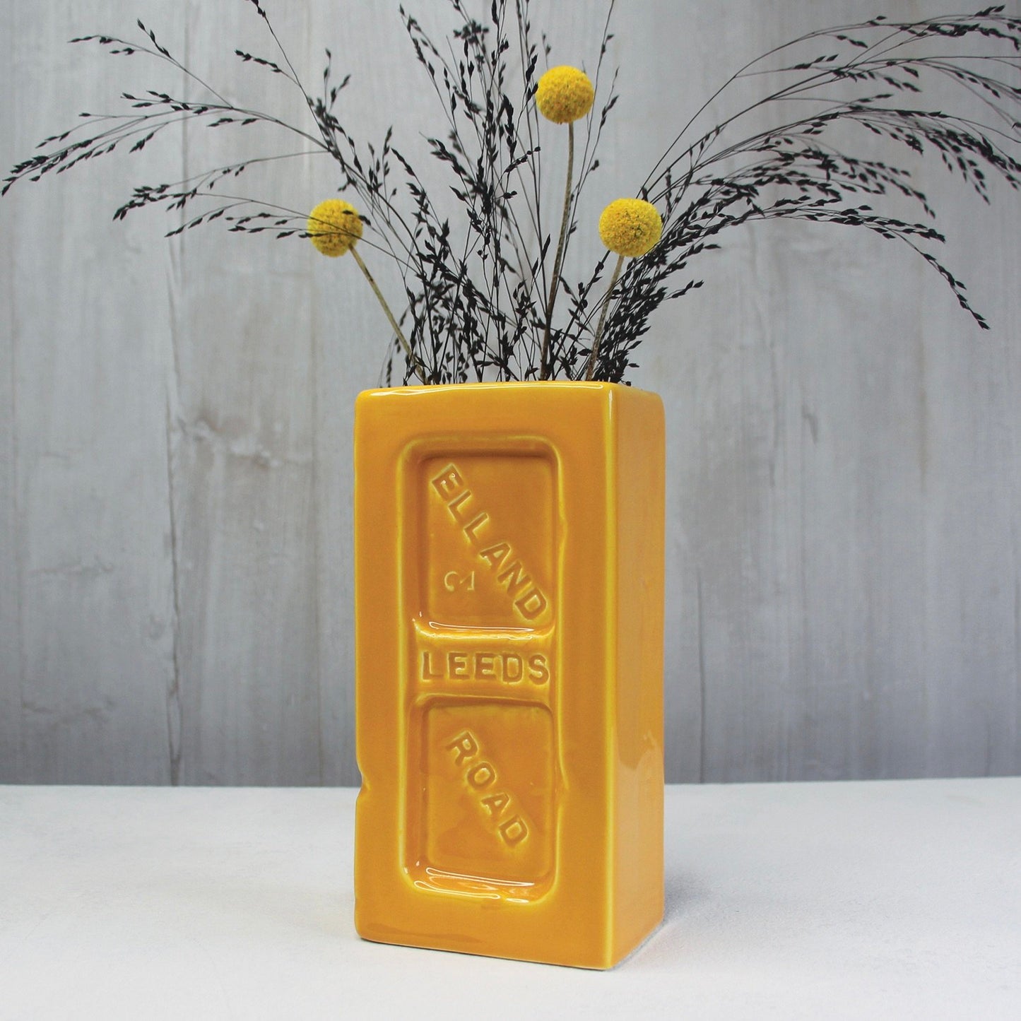 Yellow brick-shaped vase with dried flowers against a grey background.