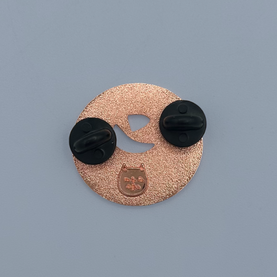 The back of a circular rose gold textured enamel pin shaped like a cat face with rubber backs on a solid gray background.
