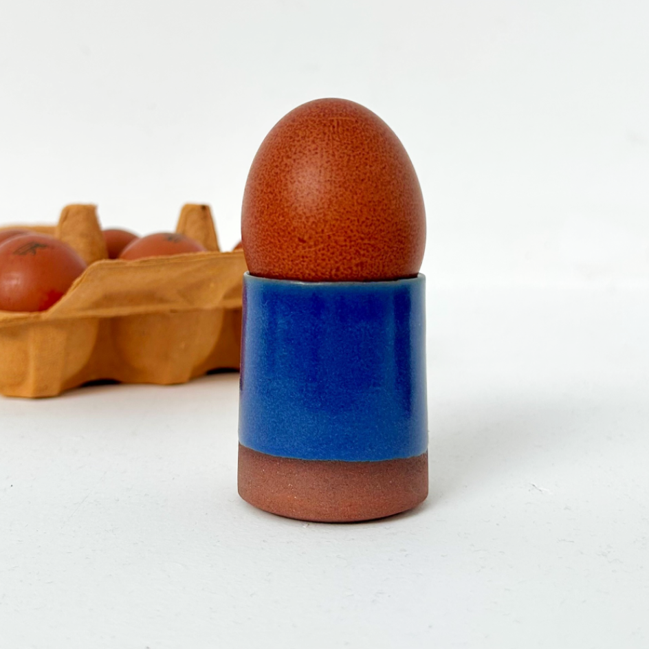 A ceramic egg cup holder with a glossy blue middle section and terracotta brown base, holding a brown speckled egg, with a blurred carton of eggs in the background on a white surface.