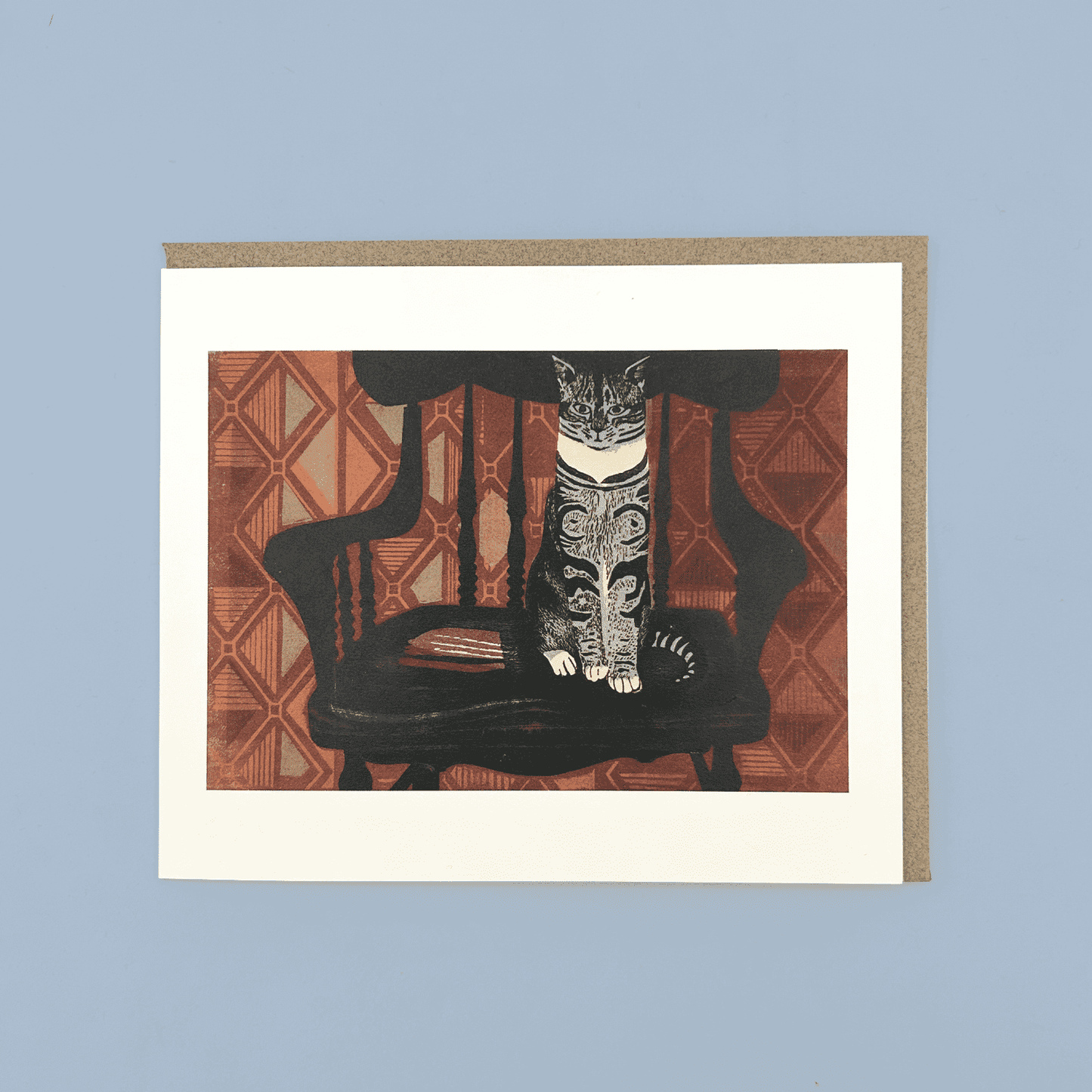 Greetings card with a linocut print of a cat sat on a chair in front of orange wallpaper. The card is on a brown envelope on