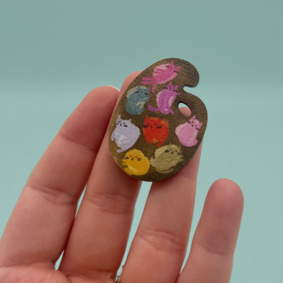 A hand holding a small wooden palette-shaped brooch decorated with colorful hand-painted cat faces, set against a solid light teal background.