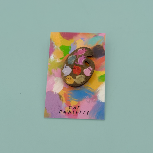 A cat-shaped painter's palette pin with multicolored paint strokes, displayed on a colorful abstract art card labeled 'CAT PAWLETTE' against a solid light teal background.