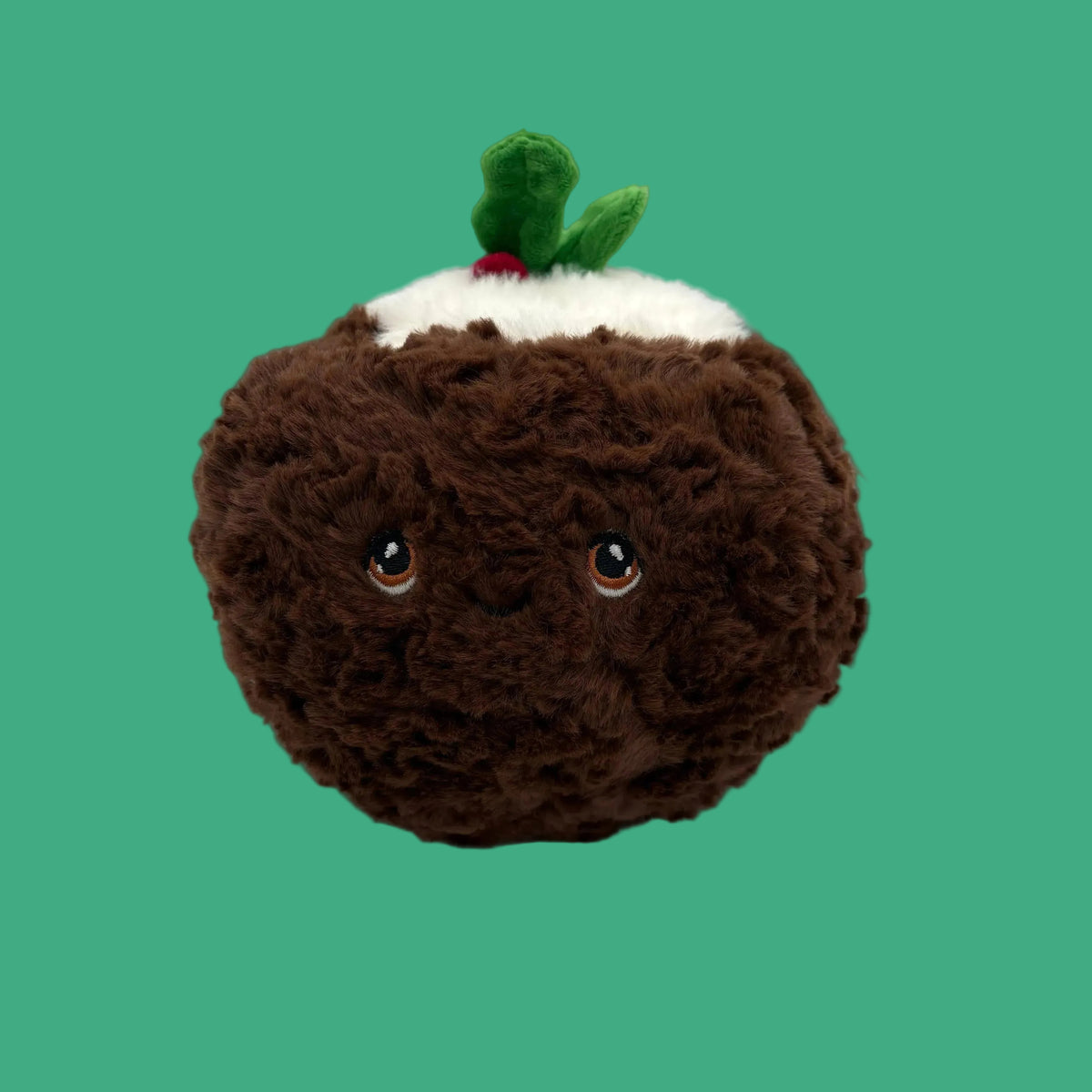 A plush pudding toy with a brown fuzzy body, white whipped cream on top, and a small green holly sprig with a red berry accent. The plush has embroidered eyes and a stitched smile, set against a neutral gray background.