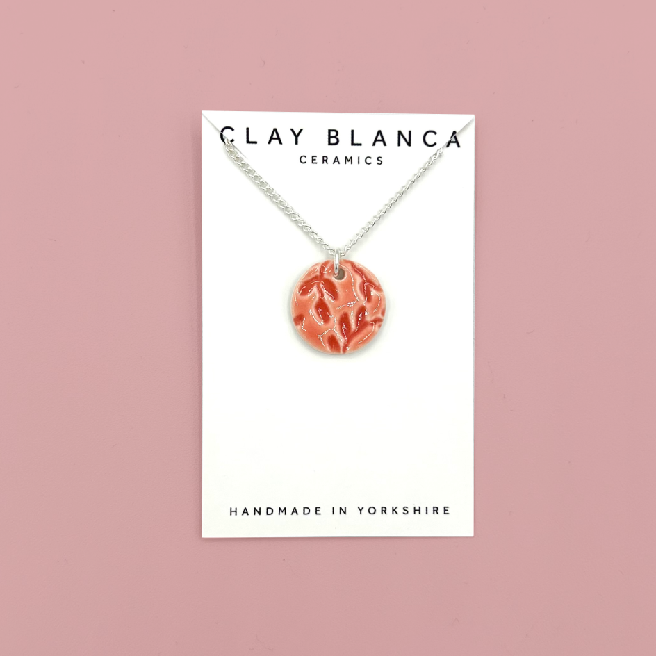 A clay pendant necklace by Clay Blanca Ceramics, featuring a circular terracotta-colored pendant with a leaf pattern, hanging from a silver chain on a white display card against a dusty pink background.