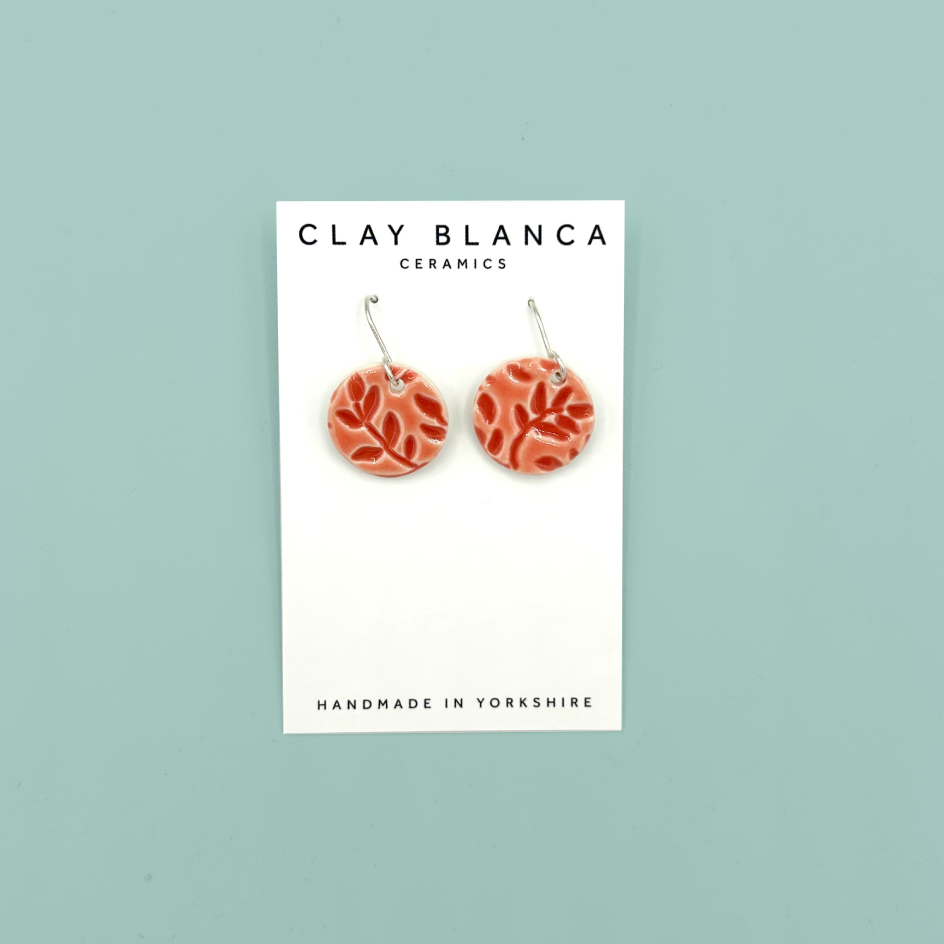 A pair of round red patterned ceramic dangle earrings on a white display card with 'CLAY BLANCA CERAMICS' printed at the top, set against a solid light teal background.