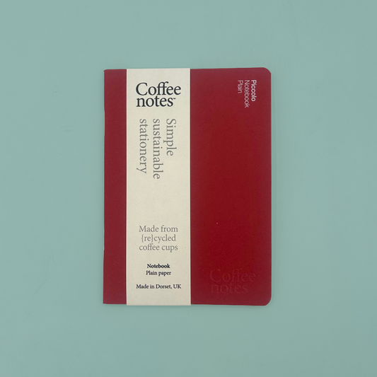 A Coffee Notes stationery notebook with a deep red cover and a cream-colored vertical label, photographed flat on a solid light teal background.