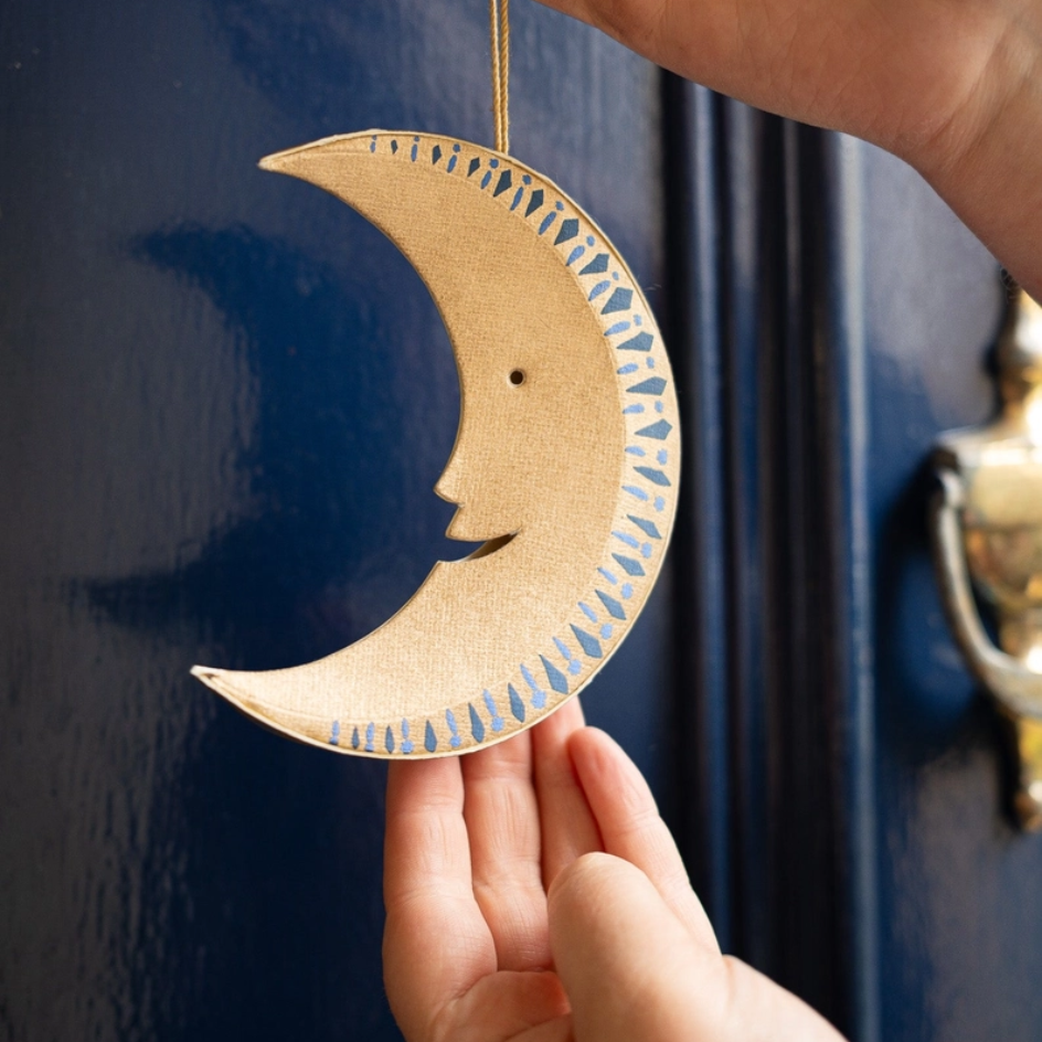 A hand holds a gold crescent moon hanging ornament in front of a dark blue door. The ornament has a cutout face and a blue patterned border.