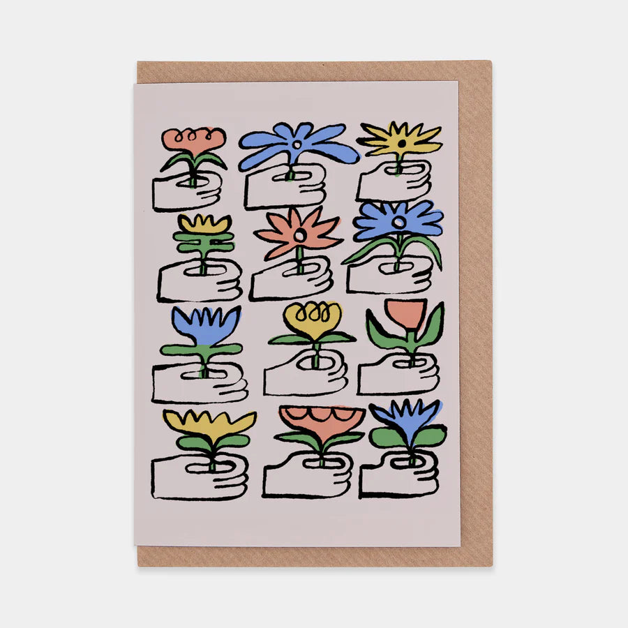 A portrait orientation greetings card with an illustration of a grid of 12 hands giving blue, red, and yellow flowers on a beige background. The card is on a brown envelope on a plain backdrop.