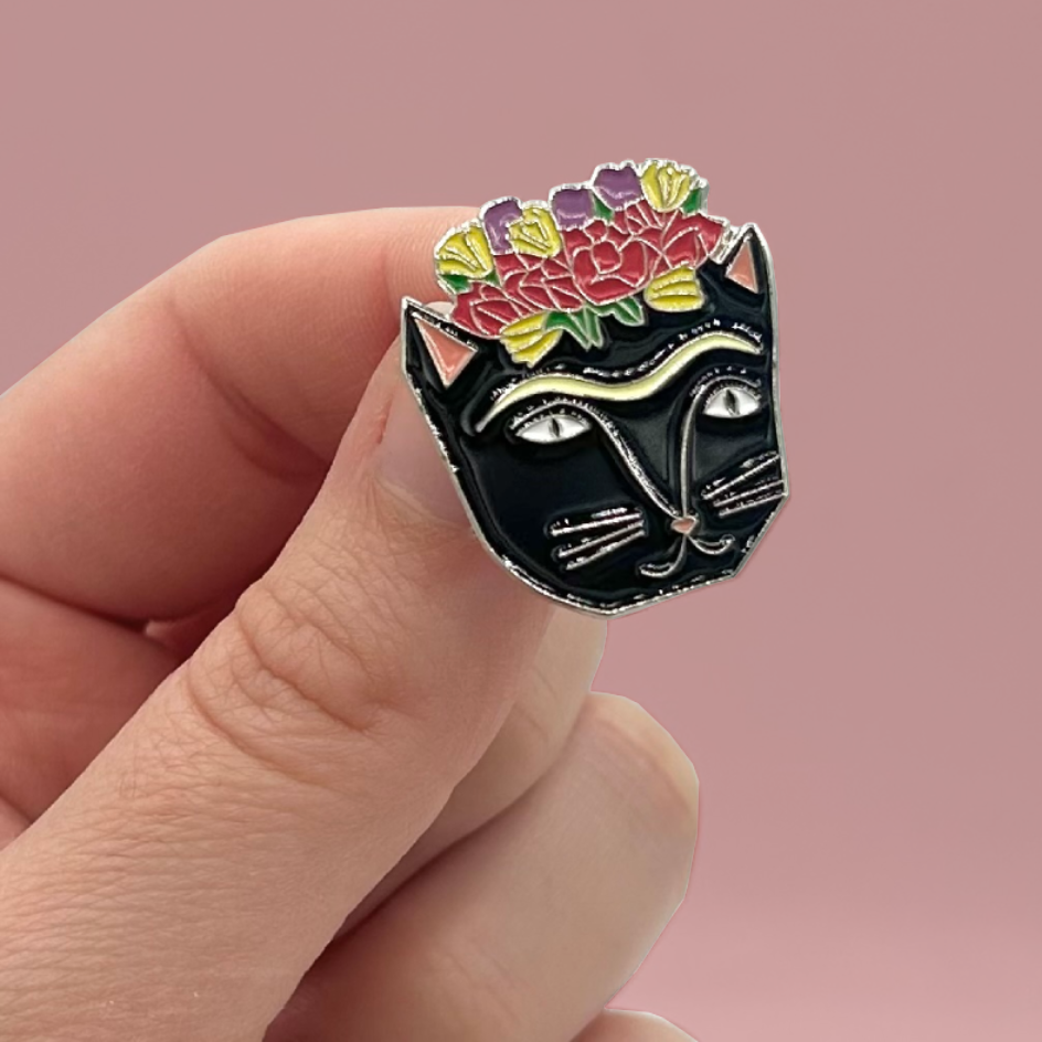 A close-up of a hand holding a black cat face enamel pin with colorful flowers on its head against a soft pink background.
