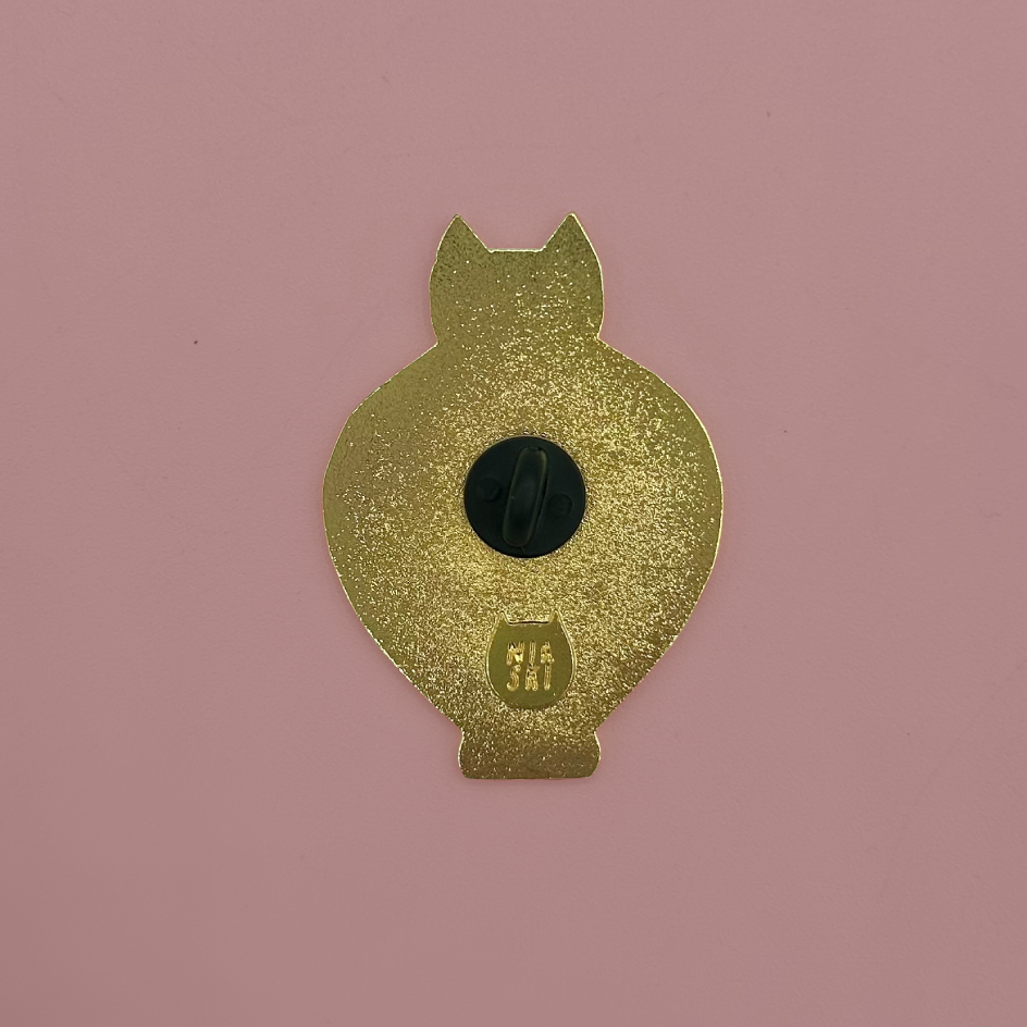 The back of a gold cat-shaped enamel pin with a black center and a small 'Niaski' stamp, shown on a solid pink background.