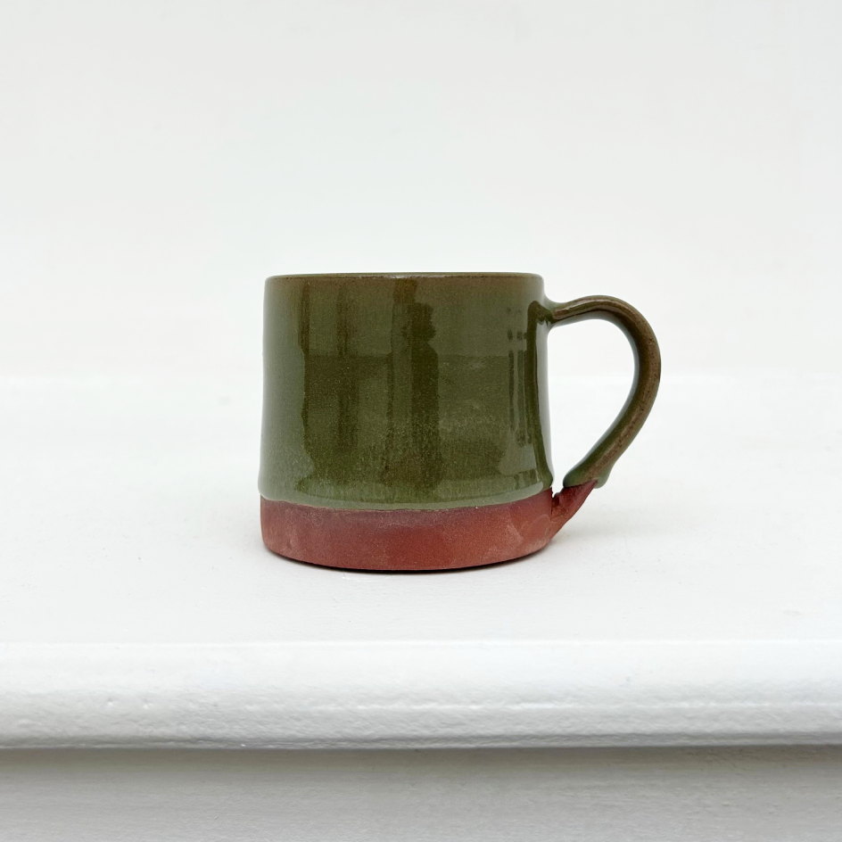 A ceramic mug with a terracotta coloured base and an olive green glossy glaze. The mug is a on a white ledge with a white wall behind.