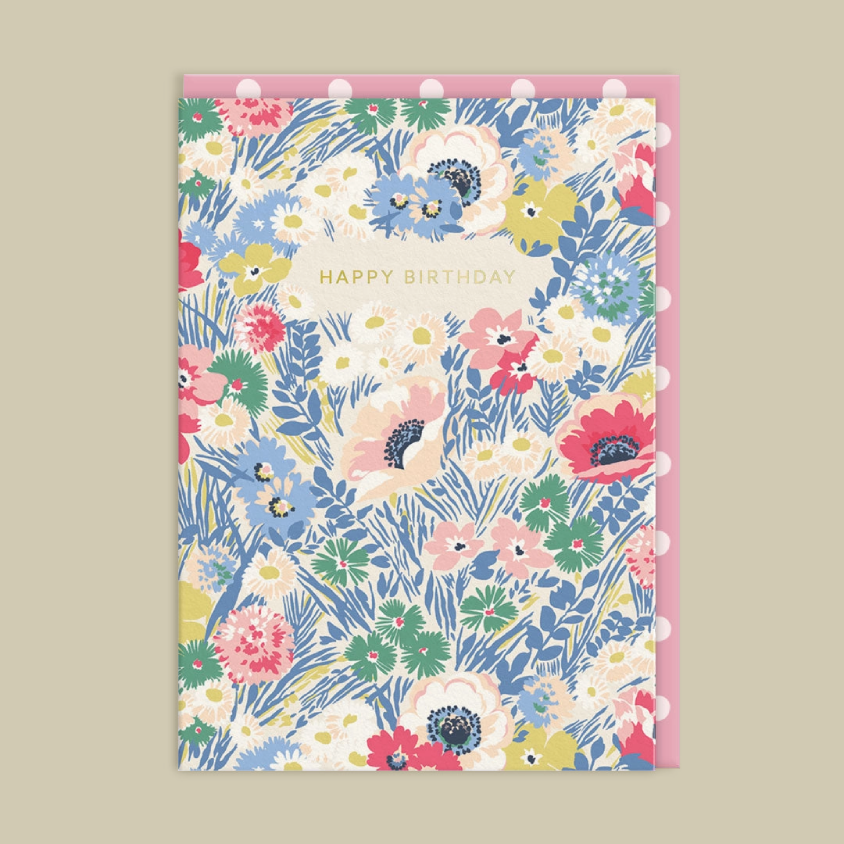A floral birthday card featuring a multicolor pattern of flowers and green leaves on a cream background with the words 'HAPPY BIRTHDAY' in gold. It is shown with a matching pink envelope on a solid beige background.