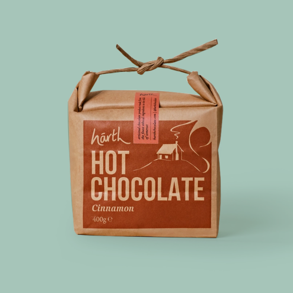 A square kraft paper package of Harth Hot Chocolate Cinnamon, featuring a brown label with white text and graphics, tied with a twine handle on a solid light green background.