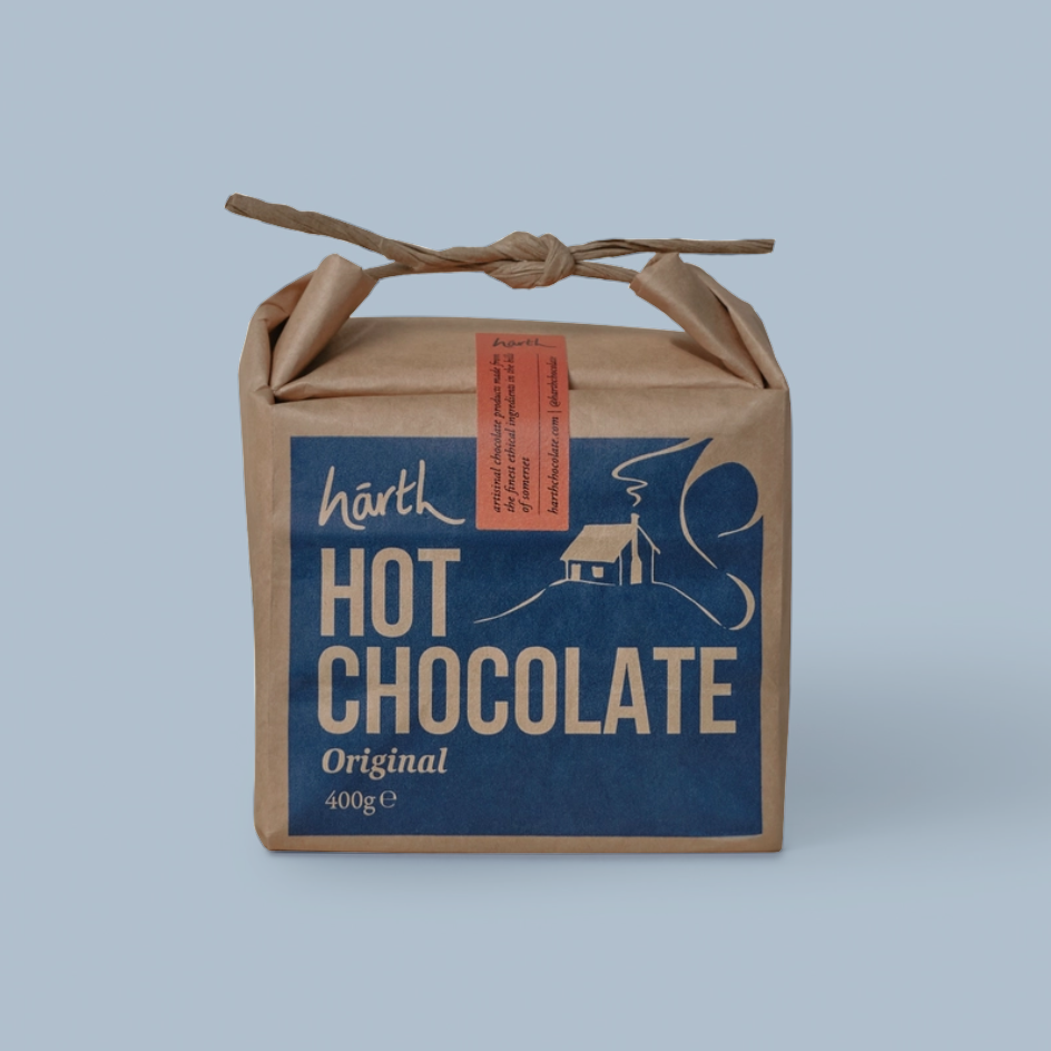 A kraft paper gift box tied with a twine handle, featuring a blue front label that reads 'harth HOT CHOCOLATE Original 400g' with a cream illustration of a house and cocoa bean. A small orange tag is attached on the side, set against a solid light blue background.