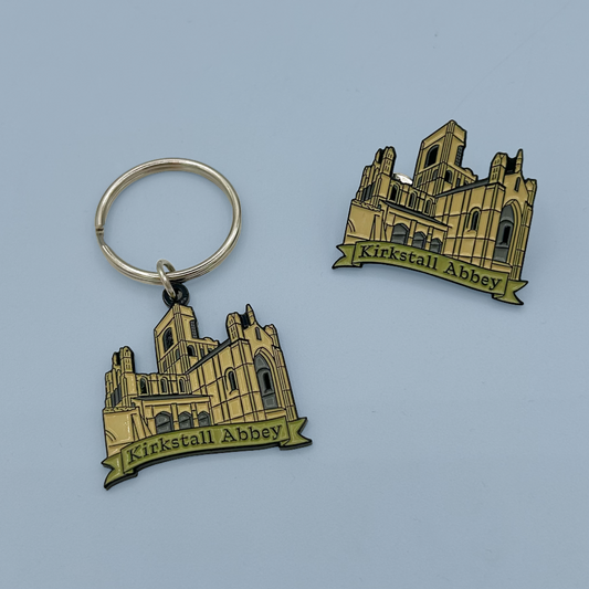 A metal keychain and matching pin featuring an embossed Kirkstall Abbey design in gold and black, displayed on a light blue background.