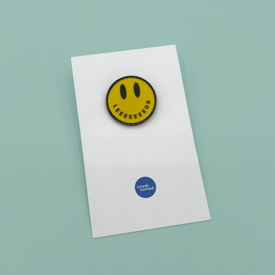 A yellow smiley face enamel pin with black eyes and the word 'LEEEEDS' in black, mounted on a white display card with a small blue 'Leeds enamel' logo near the bottom. The card is photographed on a light mint green background.