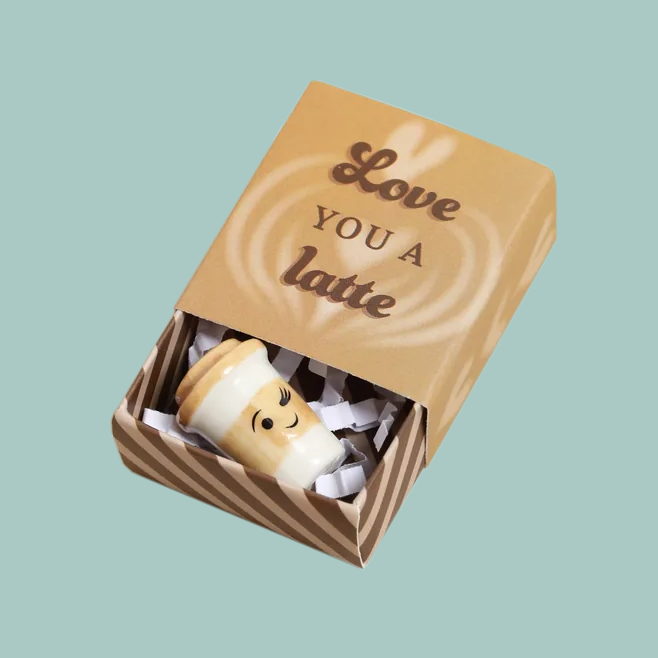 A brown cardboard matchbox with 'love you a latte' in cursive font, opened halfway, to show a ceramic latte cup with a winking face.