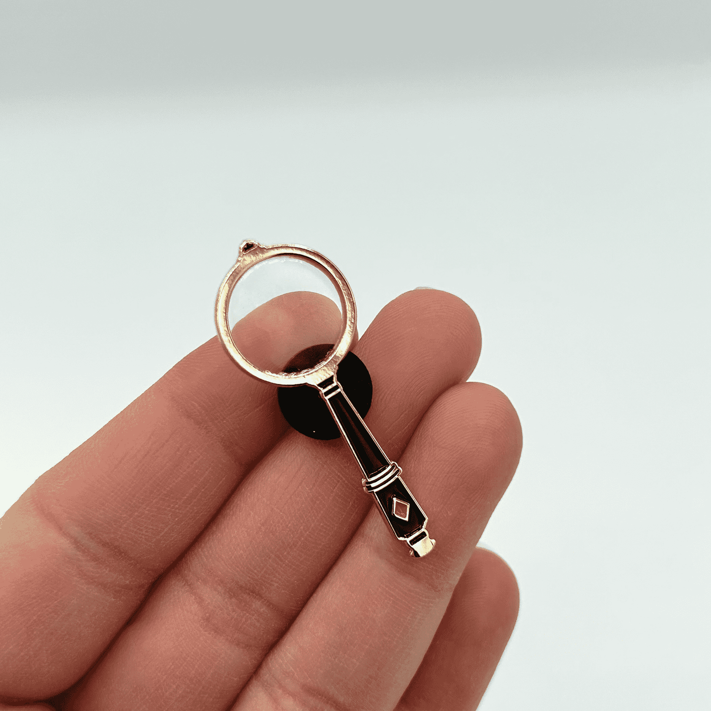 Magnifying glass pin badge held on a hand against a light background.