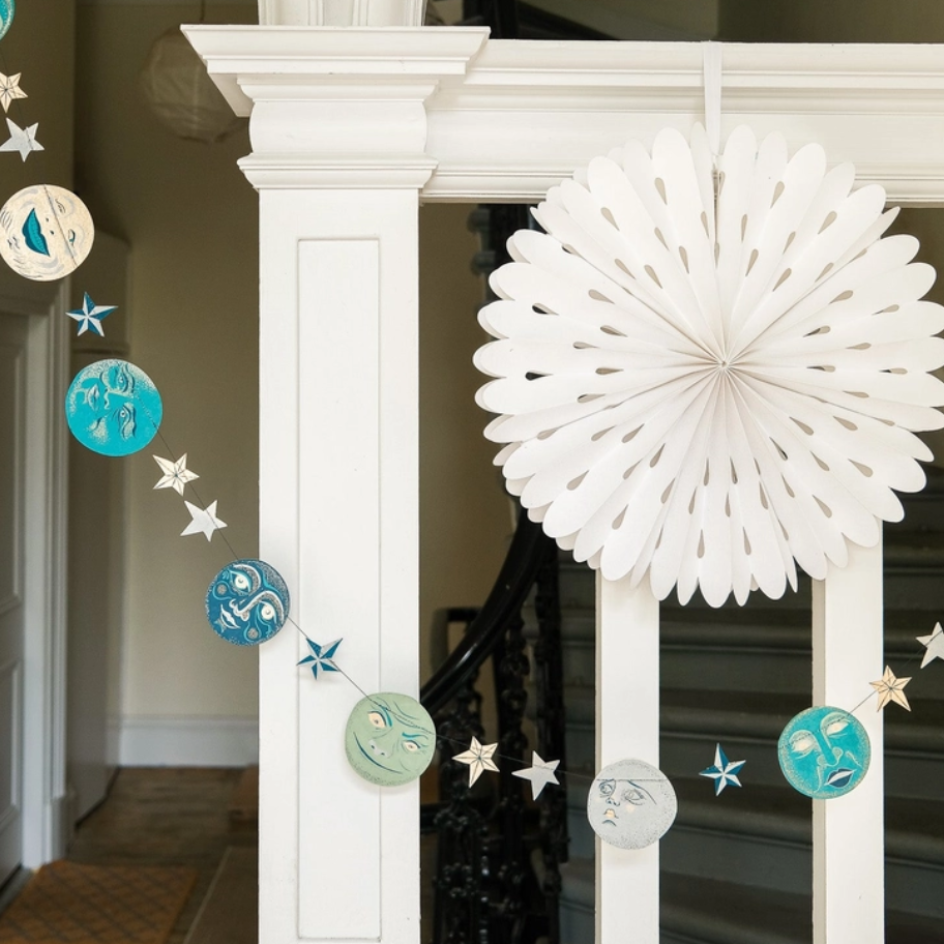 Celestial-themed wall hanging decoration featuring a large white paper fan and a matching banner with moons and stars in blue, teal, and white, displayed on a white staircase railing.