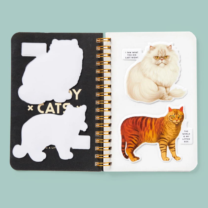 A spiral-bound sticker book or journal shown open against a pale blue background. The left page is black with white cat silhouettes and gold text reading 'COZY CATS'; the right page has two cat stickers, one cream-colored and one orange tabby, with small text bubbles.