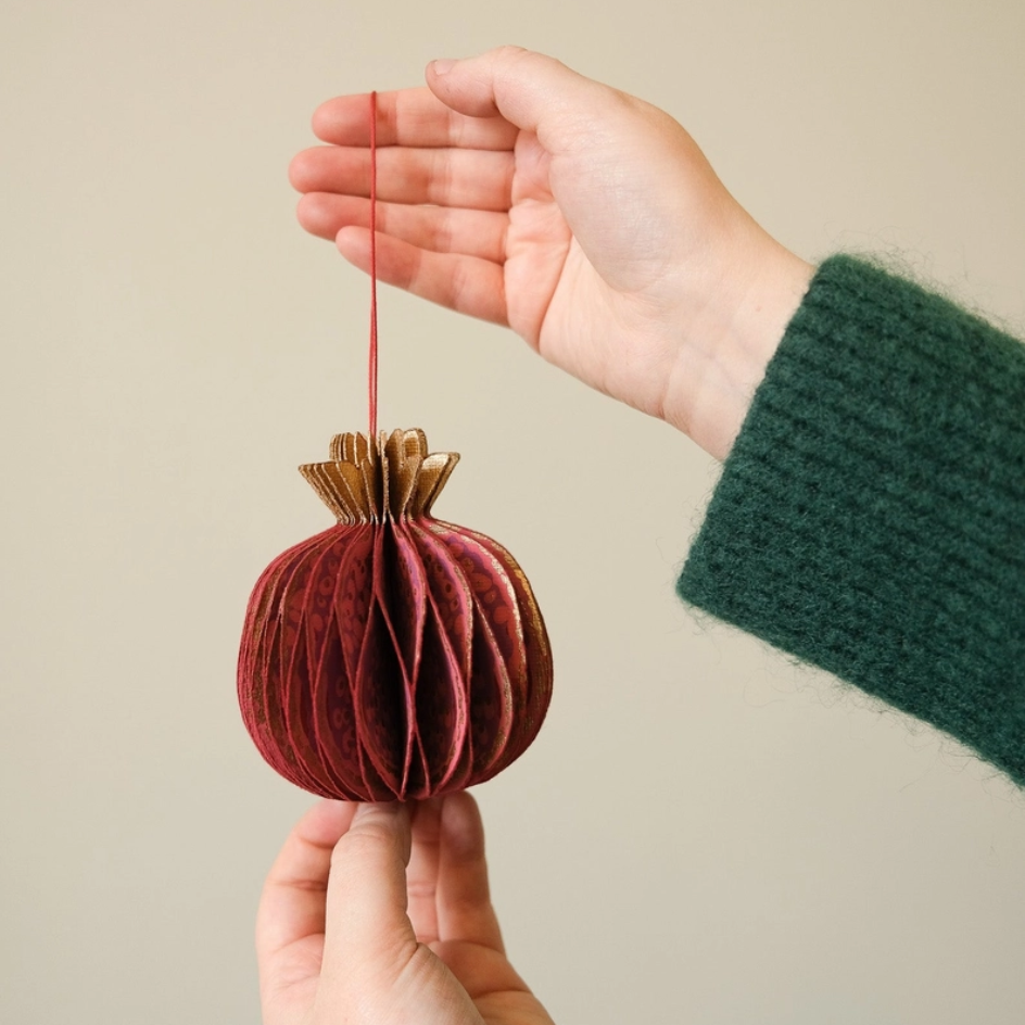 A person holds a small red paper honeycomb ornament with a gold top and a red hanging string against a plain beige background.
