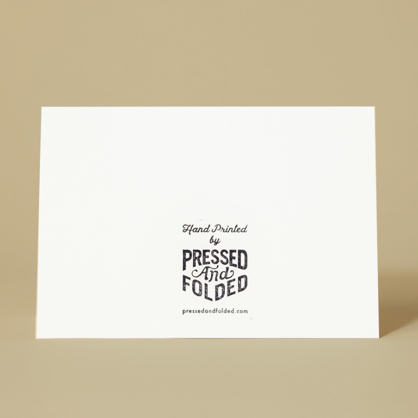 The back of a white rectangular card standing upright against a warm beige background. The center features black text reading 'Hand Printed by PRESSED And FOLDED' with the website 'pressedandfolded.