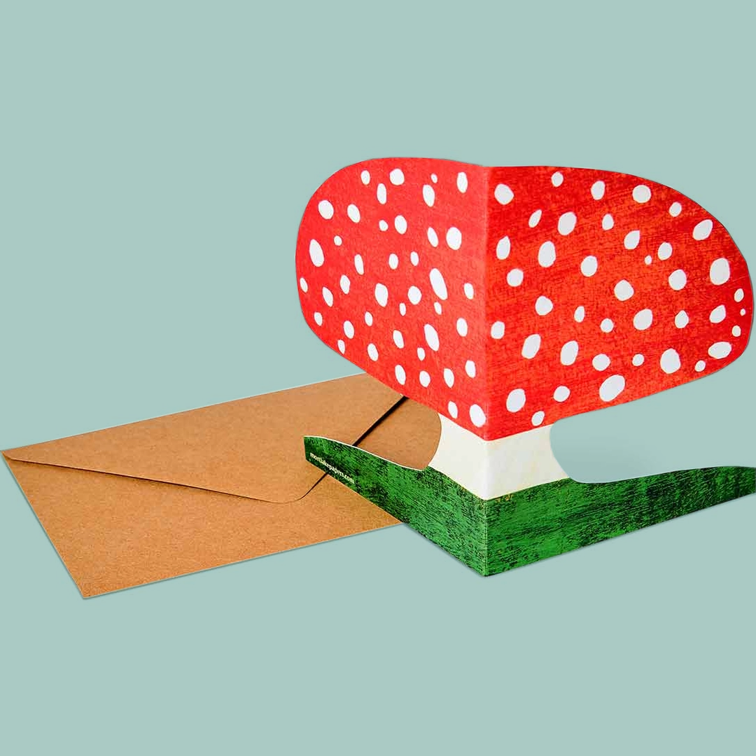 Greetings card of a mushroom with red cap and white spots, with a brown envelope on a light blue background