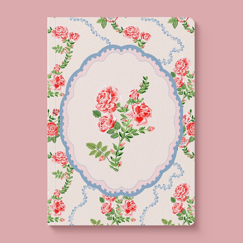 A hardcover notebook with a vintage floral design, featuring pink roses and green leaves around an ornate blue and pink oval frame on a cream background, set against a solid dusty pink backdrop.