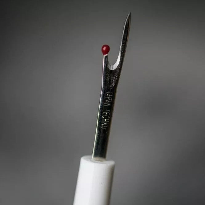Close up of a white seam ripper, on a grey background.