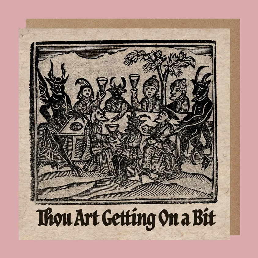 A square greetings card with an illustration of medieval figures around a table, with the text 'Thou Art Getting On a Bit' below. The card is shown against a kraft paper enveloped on a pink background.