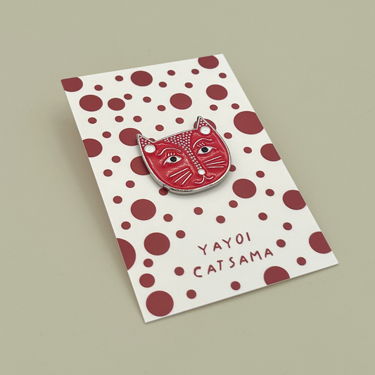 A red and white cat-face enamel pin with white polka dots and the text 'YAYOI CATSAMA' on a white backing card with red polka dots, photographed on a plain beige background.