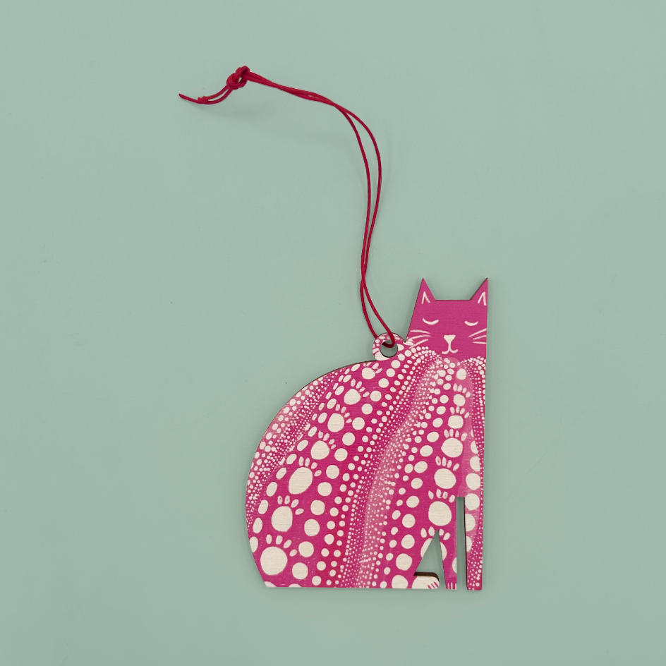 A pink cat-shaped ornament with a red hanging string, featuring a white polka dot pattern on the body and a solid pink head, set against a solid light green background.
