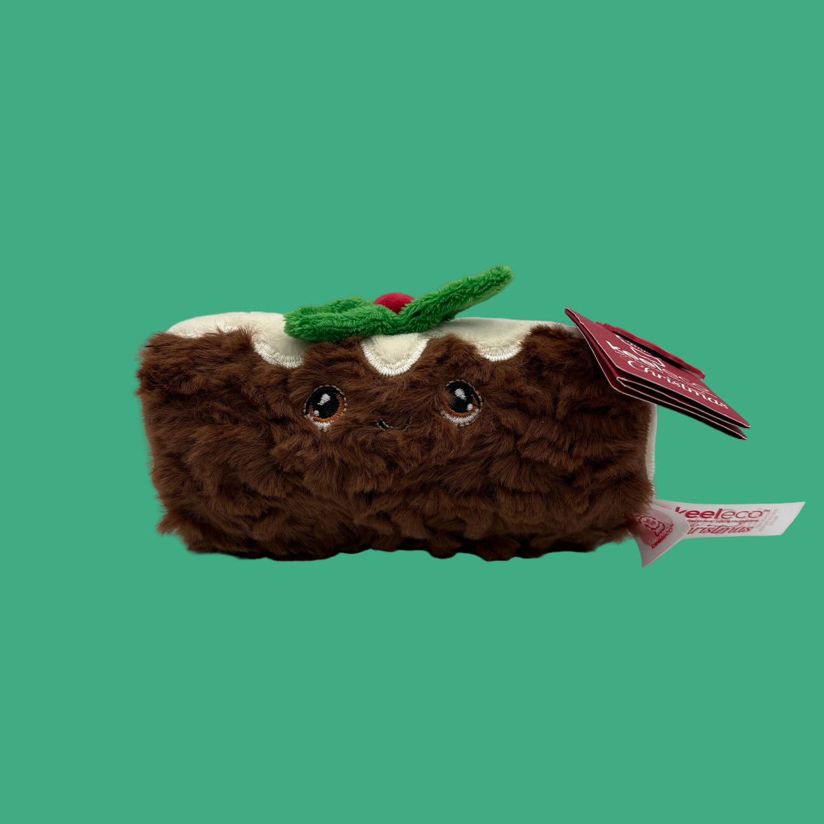 A brown plush toy shaped like a slice of cake or pudding, with white icing, a green holly accent, and embroidered eyes. A red hangtag is attached on the right side, photographed on a clean light gray studio background.