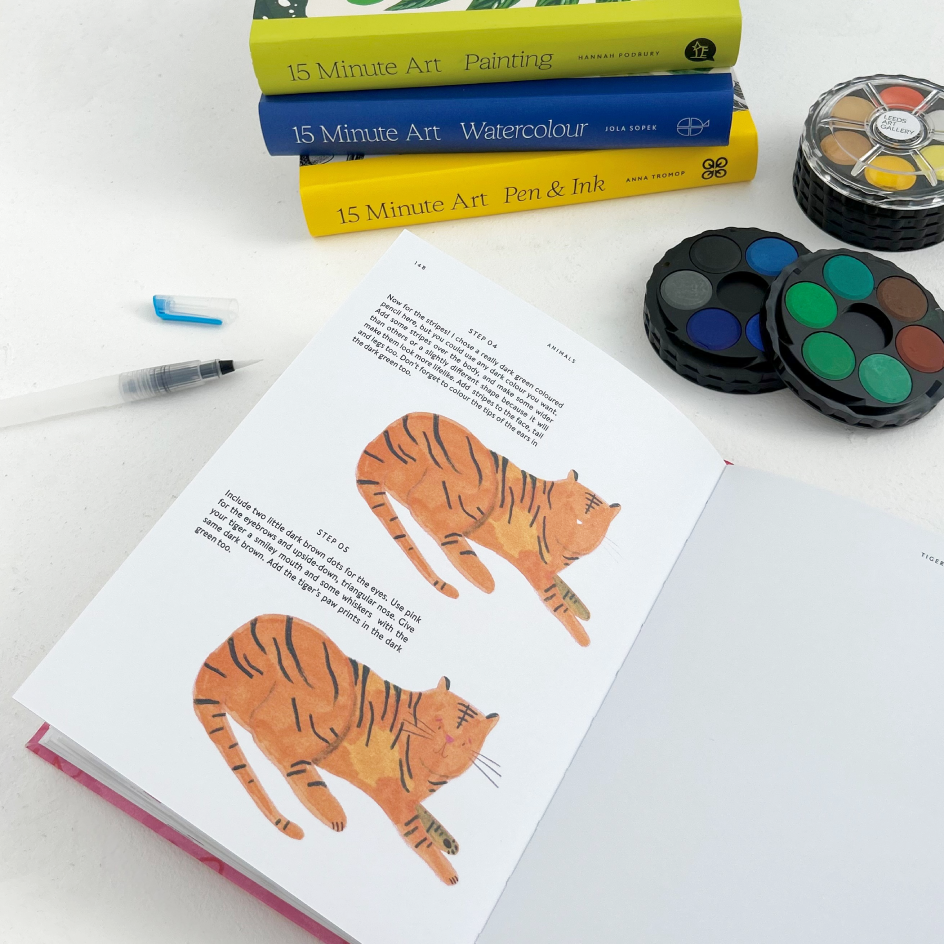 An open art instruction book showing watercolor tiger illustrations and text, arranged on a white surface with paint palettes, a pen, and stacked 15 Minute Art books in the background.