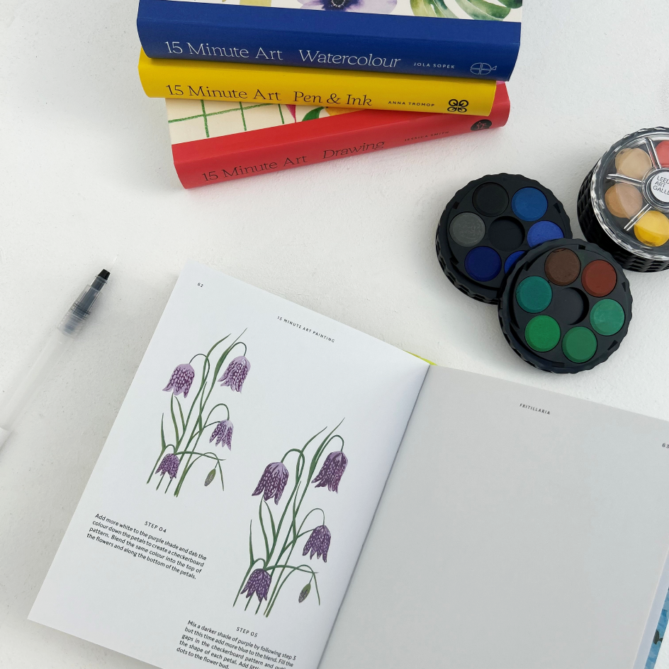 An open art instruction book showing purple flower illustrations, arranged on a white surface with stacked 15 Minute Art books, two round watercolor palettes, and a pen.