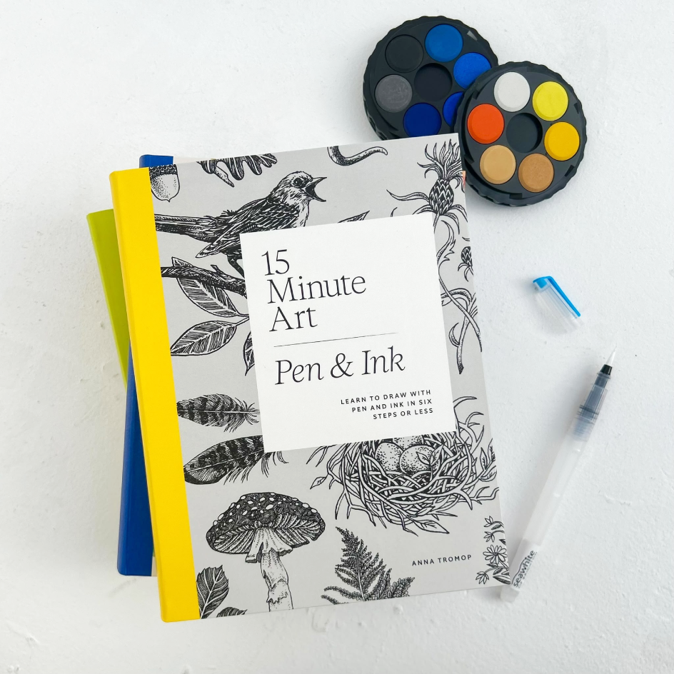 A book titled "15 Minute Art: Pen & Ink" lies on a white surface, surrounded by art supplies including a round watercolor palette and a paintbrush.
