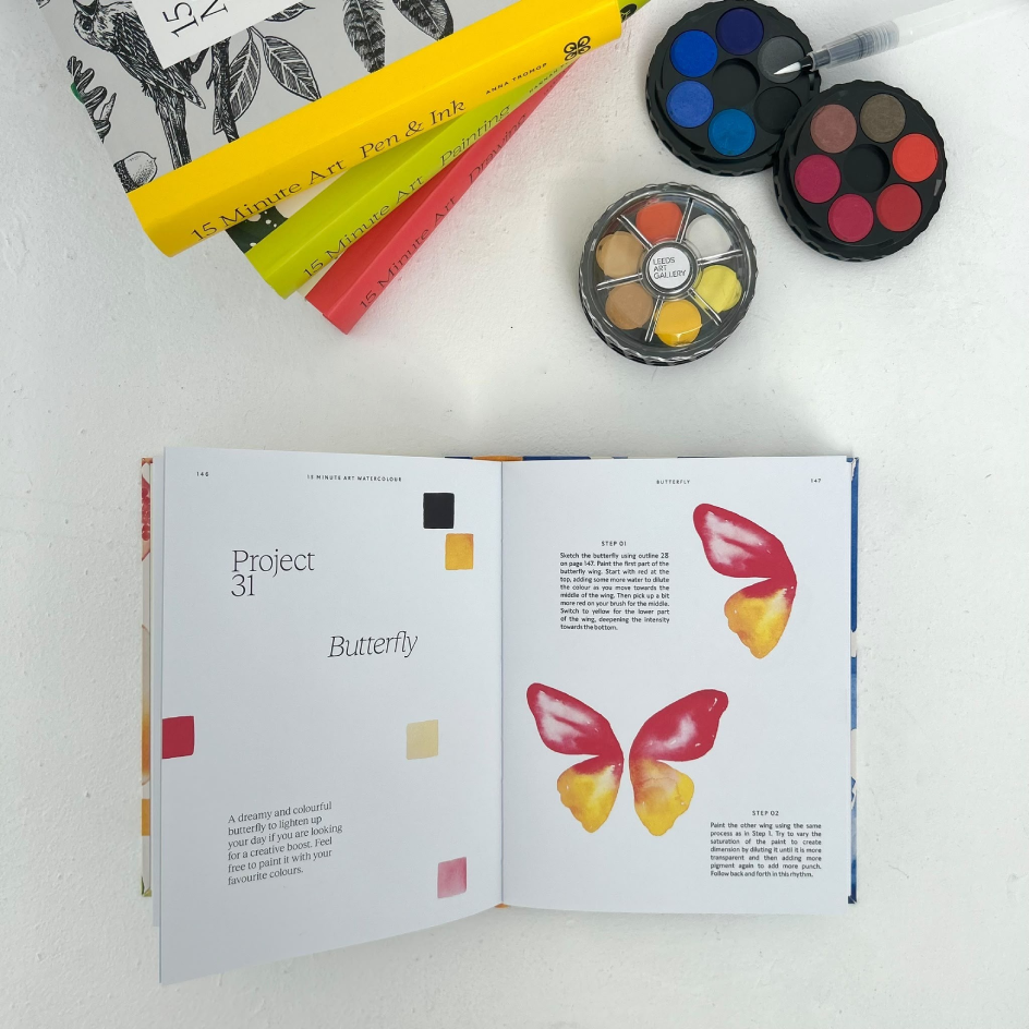 An open art book spread showing butterfly color studies, styled on a white surface with paint palettes and brushes