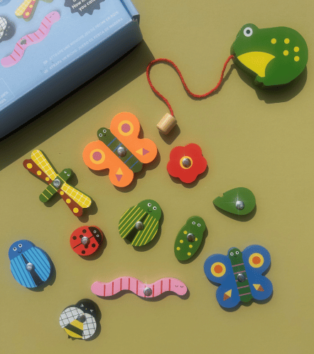 A set of colorful wooden insect and worm puzzle pieces with red hanging strings, arranged on a muted green surface next to a blue box.