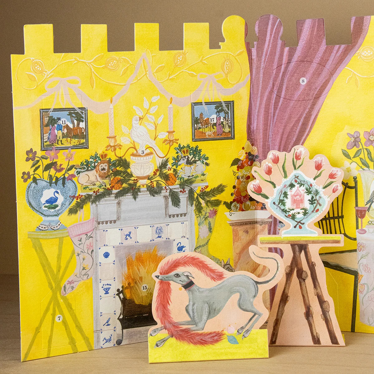 A colourful illustrated advent calendar featuring a yellow fireplace scene with flowers, stockings, framed pictures, and layered paper cutout figures including a gray dog with pink fur and a vase on a wooden stand.