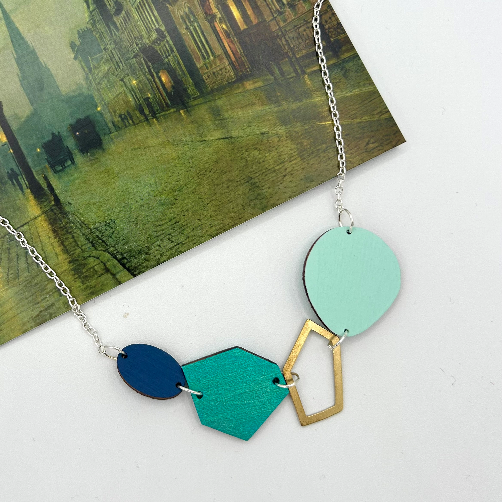 A mint and navy geometric necklace on a silver chain. The necklace is on a postacrd of Atkinson Grimshaw's 'Park Row.'