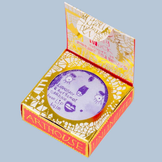 An open Arthouse Unlimited lip balm box in gold and red packaging, showing a round purple label with handwritten text inside, set against a solid light blue background.