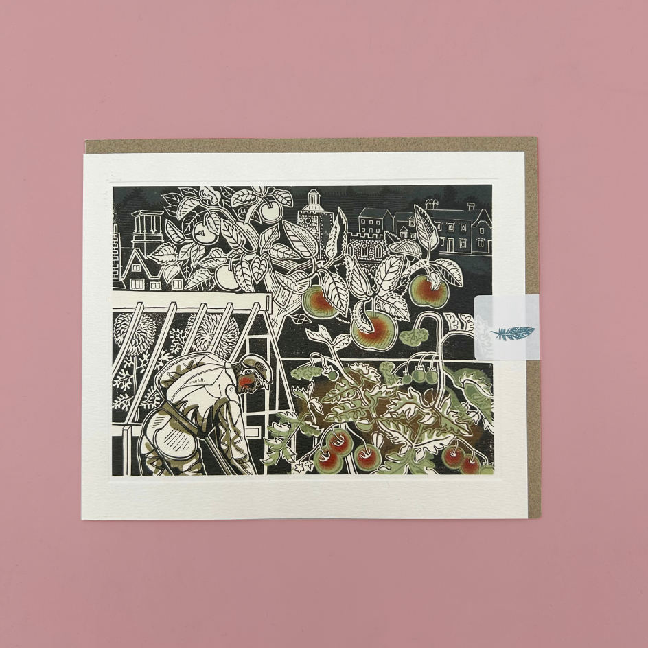 A matted greeting card featuring a black-and-white linocut-style illustration of fruit trees and a gardener, with small muted red and green accents on the fruit. The card is photographed flat against a dusty pink background, with a kraft envelope partially visible behind it.