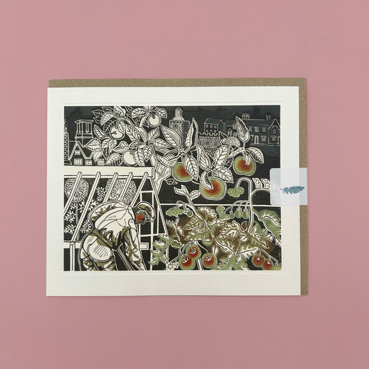 A matted greeting card featuring a black-and-white linocut-style illustration of fruit trees and a gardener, with small muted red and green accents on the fruit. The card is photographed flat against a dusty pink background, with a kraft envelope partially visible behind it.