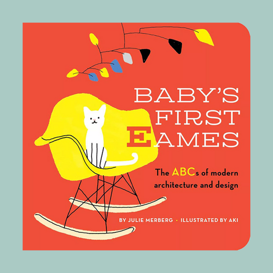Book cover for 'Baby's First Ames: The ABCs of modern architecture and design' by Julie Merberg, illustrated by Aki. The cover features a bold coral-red background with a yellow rocking chair, a white cat, and modern abstract tree branches in yellow, blue, black, and white.