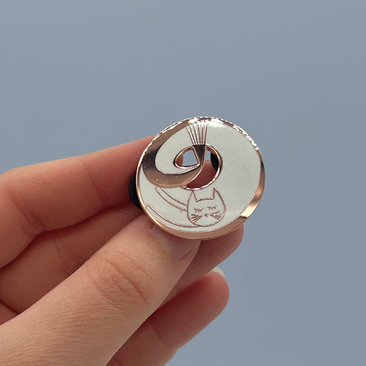 A hand holding a pin badge of a white cat with rose gold details. The background is a blue-grey.