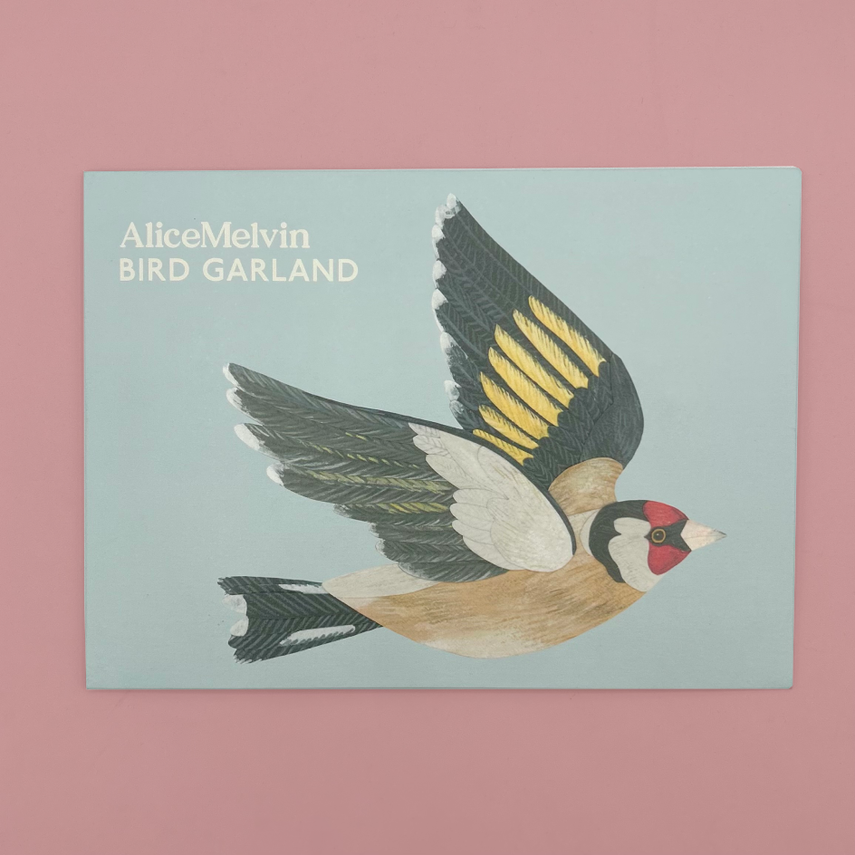 Packaging featuring an illustrated goldfinch in flight on a pale blue background, with the text 'AliceMelvin BIRD GARLAND' in the upper left. The packet is photographed against a solid pink background.