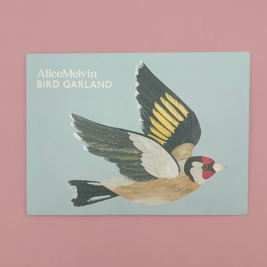 Packaging featuring an illustrated goldfinch in flight on a pale blue background, with the text 'AliceMelvin BIRD GARLAND' in the upper left. The packet is photographed against a solid pink background.
