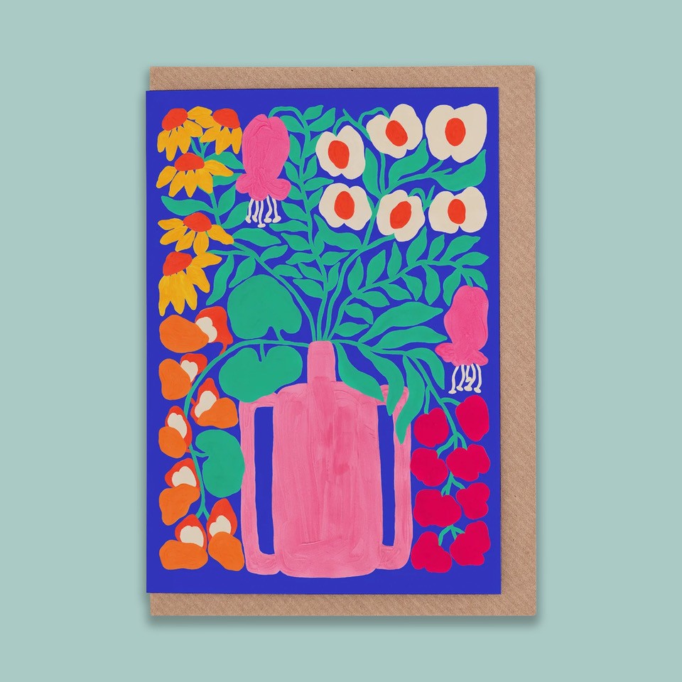 A portrait orientation greetings card with an illustration of a pink vase with flowers on a blue background. The card is on a brown envelope on a plain backdrop. 