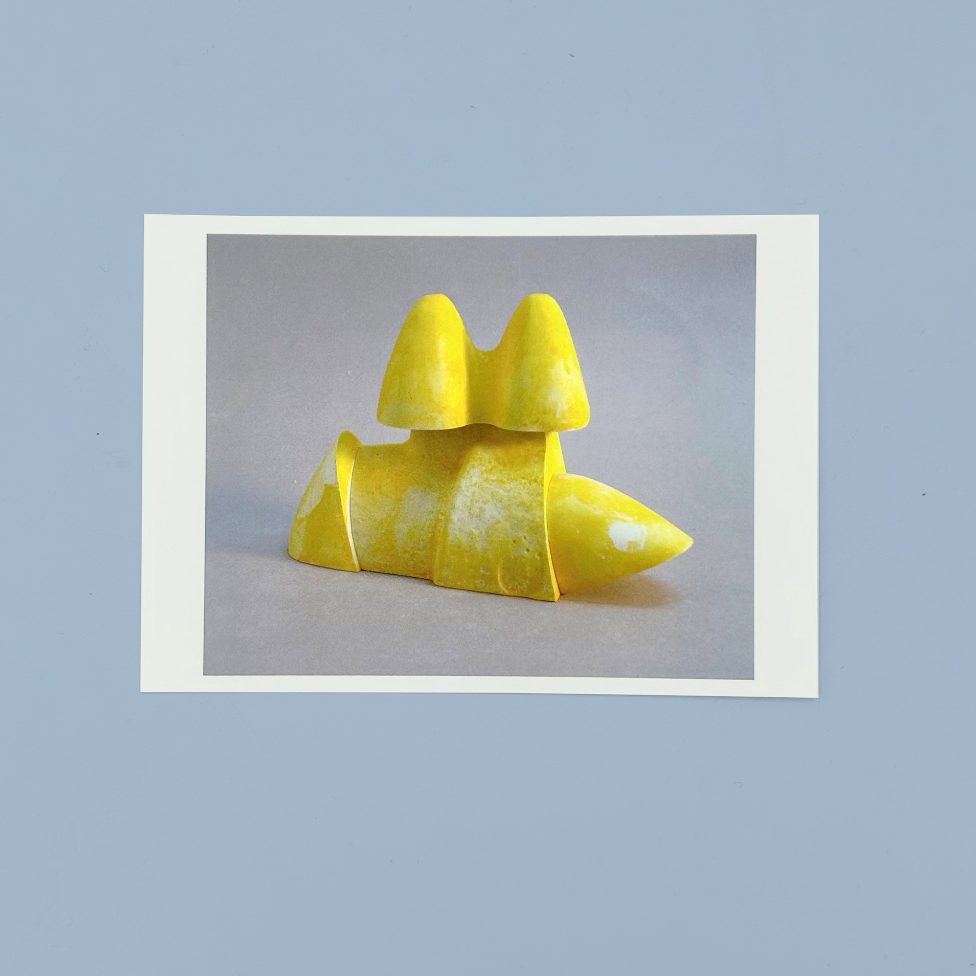 Postcard of a yellow sculpture on a gray background. The postcard is on a blue background.